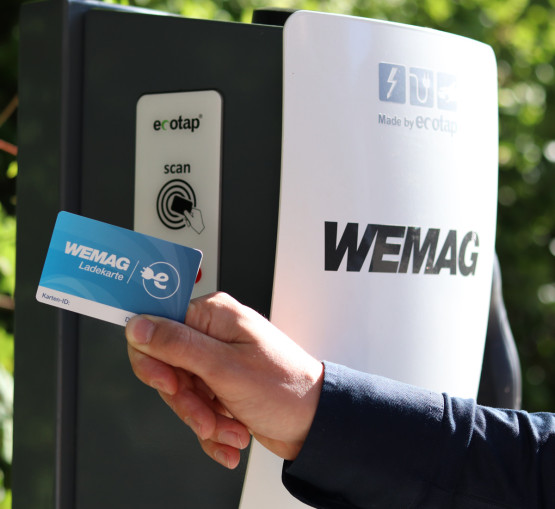 Eine Hand hält eine WEMAG Ladekarte vor eine Ladestation für Elektroautos. Die Station ist teilweise im Schatten, mit grünen Blättern im Hintergrund, betont durch Sonnenlicht. Der Fokus liegt auf der Karte und der Scan-Bereich der Station, die die einfache und bequeme Nutzung der Ladeinfrastruktur für Elektroautos durch WEMAG-Kunden zeigt.