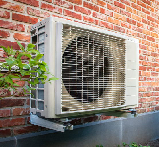 Effiziente Wärmepumpe - nachhaltiges Heizen mit WEMAG Eine Außeneinheit einer Wärmepumpe montiert an einer roten Backsteinwand neben einem grünen Strauch, demonstriert eine umweltfreundliche Heizungslösung für Wohngebäude.