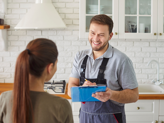 Freundlicher Monteur im Hintergrund mit blauer Mappe in der Hand, lächelt freundlich der Kundin zu.