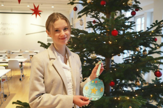 Junge Frau mit rundem Wunschzettel steht vor einem Weihnachtsbaum mit roten Kugeln.