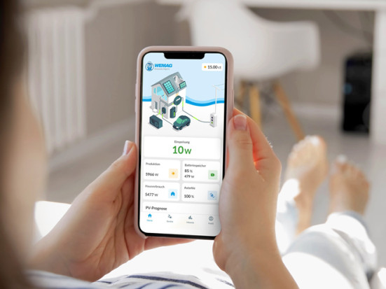 Hände halten ein Smartphone mit einer Energie-App, die Stromerzeugung und -verbrauch eines Hauses anzeigt; im Hintergrund liegt eine Person entspannt auf einem Sofa in einem hellen Wohnzimmer.