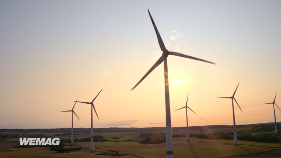 Windkraftanlagen in einer Landschaft bei Sonnenuntergang. Felder und Wiesen im Vordergrund sichtbar.