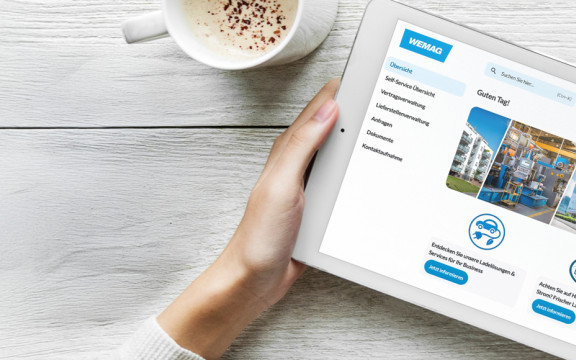 Tablet mit geöffnetem WEMAG-Geschäftskundenportal auf einem hellen Holztisch. Auf dem Bildschirm sind Menübereiche wie Vertrags-, Lieferstellen- und Dokumentenverwaltung zu sehen. Neben dem Tablet stehen eine Kaffeetasse.