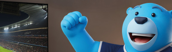Links ein Fernseher mit hell beleuchtetem Fußballstadion voller Fans während eines Spiels, rechts ein fröhlicher blauer Cartoon-Bär im Sporttrikot mit erhobener Faust.