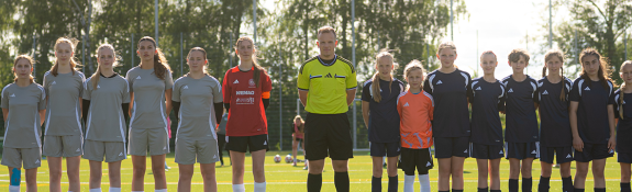 Jugendfußballteams stehen gemeinsam mit einer Schiedsrichterperson auf einem Kunstrasenplatz.