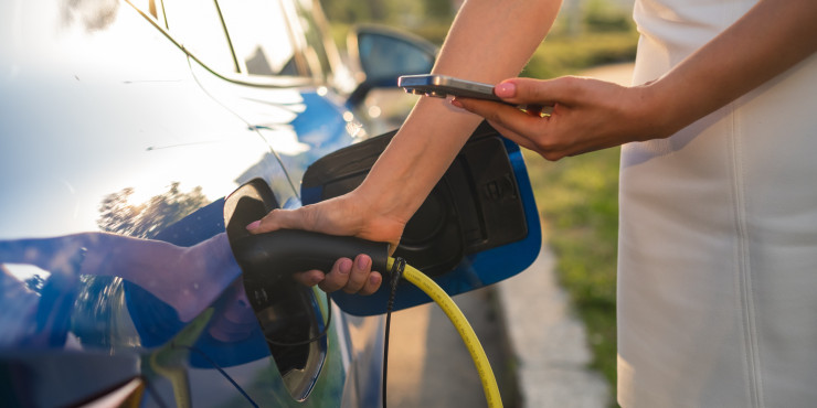 Eine Frau in einem weißen Kleid lädt ihr Elektroauto an einer Ladestation auf. Sie hält das Ladekabel in einer Hand und bedient mit der anderen ihr Smartphone. Das Auto ist blau und spiegelt die Umgebung. Die Szene spielt sich draußen an einem sonnigen Tag mit grüner Umgebung ab.