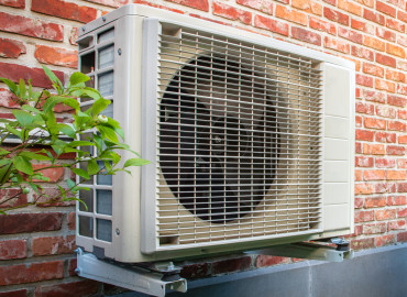 Eine Außeneinheit einer Wärmepumpe montiert an einer roten Backsteinwand neben einem grünen Strauch, demonstriert eine umweltfreundliche Heizungslösung für Wohngebäude.