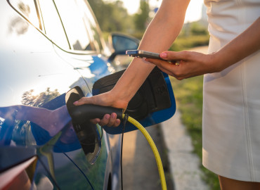Eine Frau in einem weißen Kleid lädt ihr Elektroauto an einer Ladestation auf. Sie hält das Ladekabel in einer Hand und bedient mit der anderen ihr Smartphone. Das Auto ist blau und spiegelt die Umgebung. Die Szene spielt sich draußen an einem sonnigen Tag mit grüner Umgebung ab.