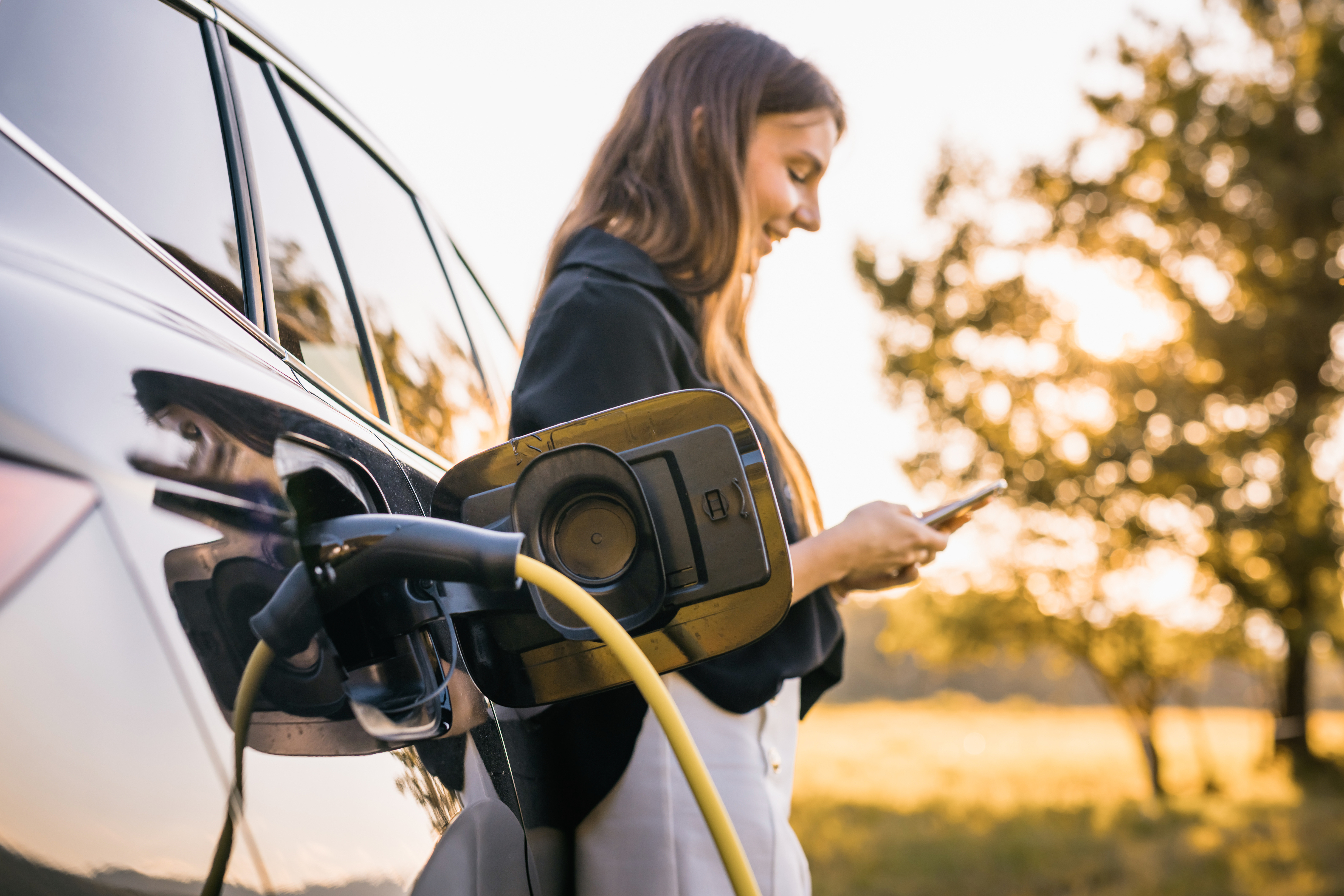 Im Vordergrund sieht man ein dunkels E-Auto, dass geladen wird. Dahimter steht eine Frau mit langen dunklen Haaren und Blickt freudig auf ihr Handy. Im Hintegrund Sonnenuntergang und Natur.