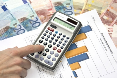 Finanzierung von Biogasanlagen - welche Möglichkeiten gibt es. Ein Taschenrechner mit der Zahl 0 und einem Finger auf den Zahlen um etwas einzutippen. Unter dem taschenrechner liegt ein Blatt mit Diagrammen und darunter Geldscheine in Höhe von 10 und 20 €.